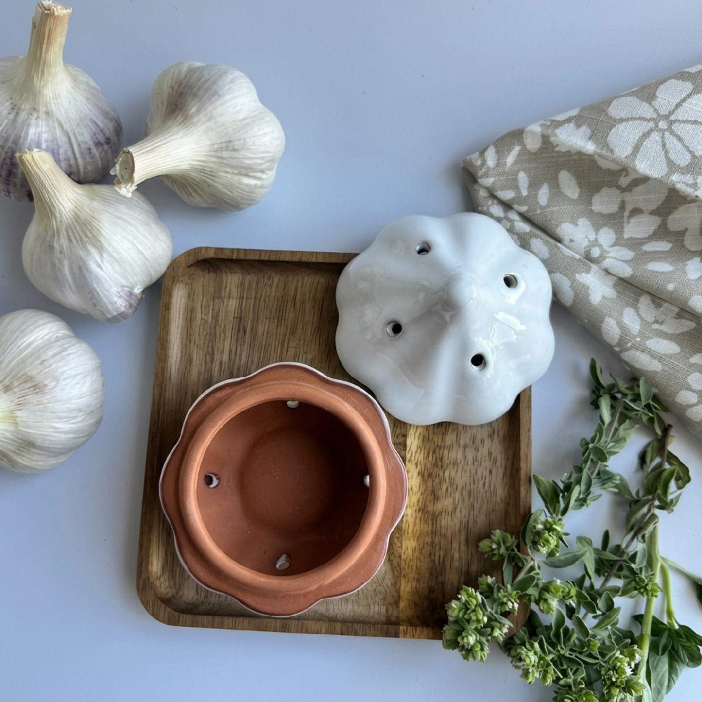 A ceramic garlic press and matching garlic holder set in terracotta and white, styled on a wooden tray with garlic bulbs, oregano, and a patterned cloth on a white background.