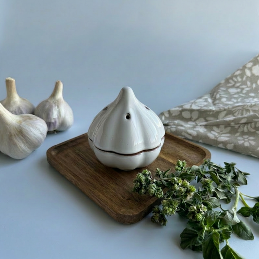 A white ceramic garlic-shaped container with a small lid sits on a wooden tray, styled with garlic bulbs, fresh herbs, and a patterned cloth against a white background.