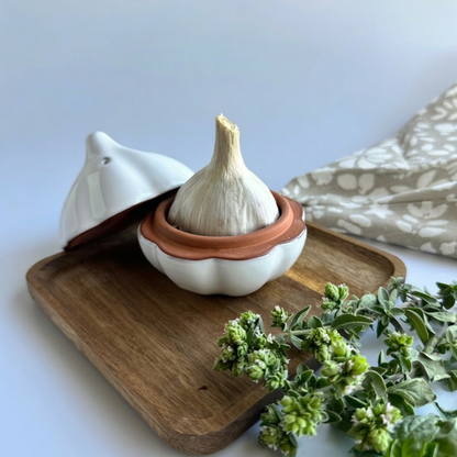 A ceramic garlic holder containing a whole garlic bulb, styled on a wooden tray with green herb sprigs and a patterned cloth in the background.