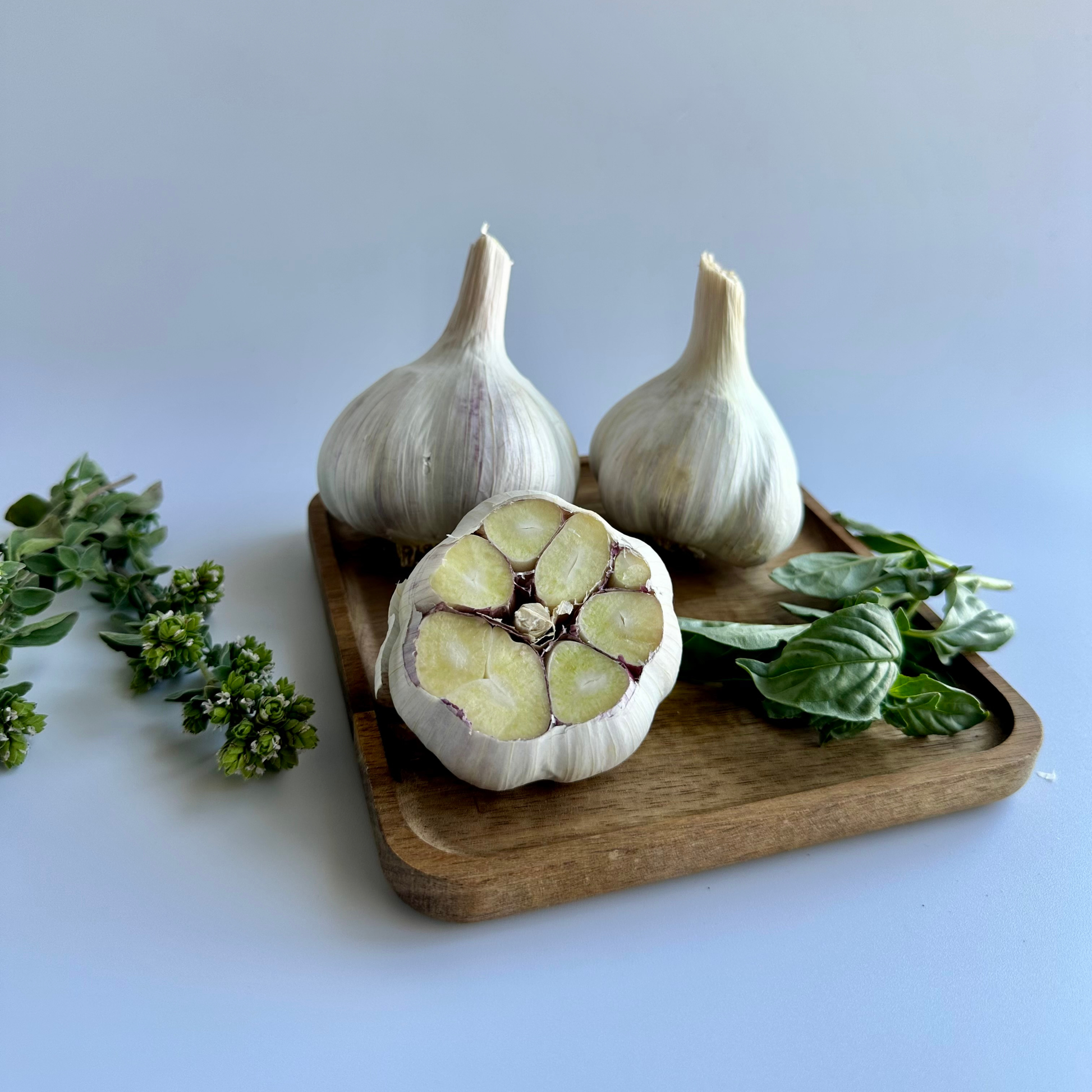 Three garlic bulbs and a cut half of a garlic bulb arranged on a small wooden tray with fresh green herbs, photographed on a white studio background.