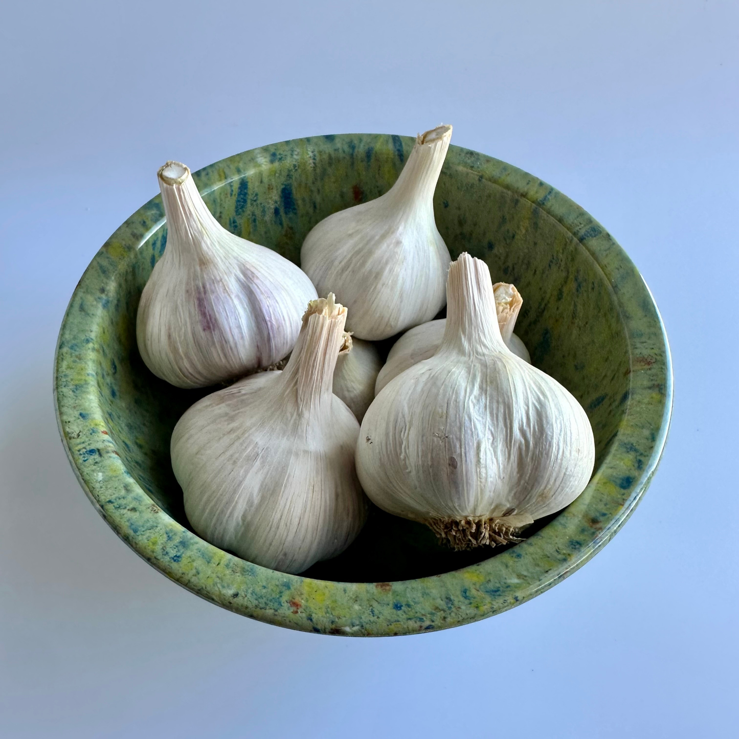 Several whole garlic bulbs resting in a round speckled green ceramic bowl against a plain light background.