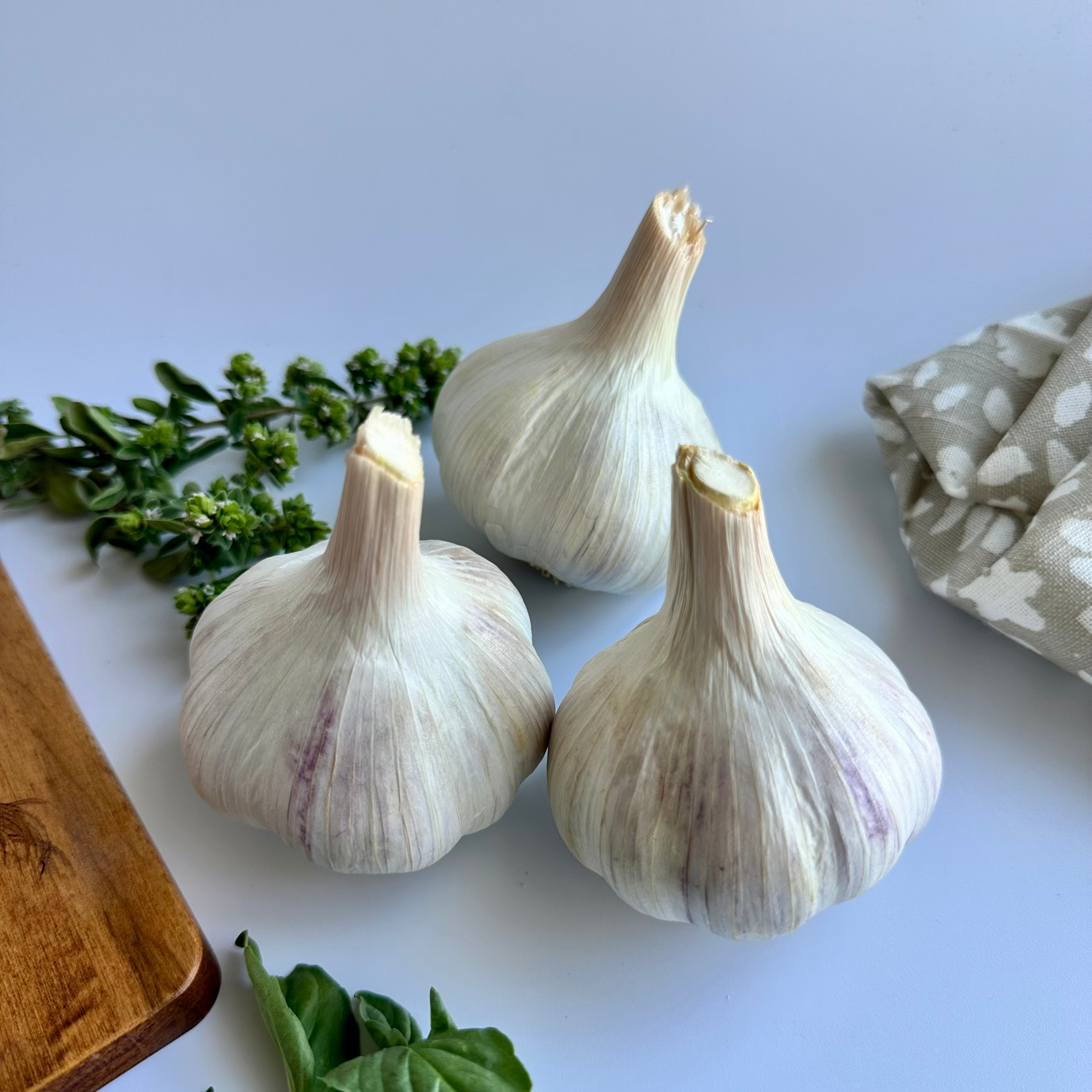 Three whole garlic bulbs arranged on a light blue surface with small green herb sprigs, a wooden cutting board corner, and a folded patterned cloth.