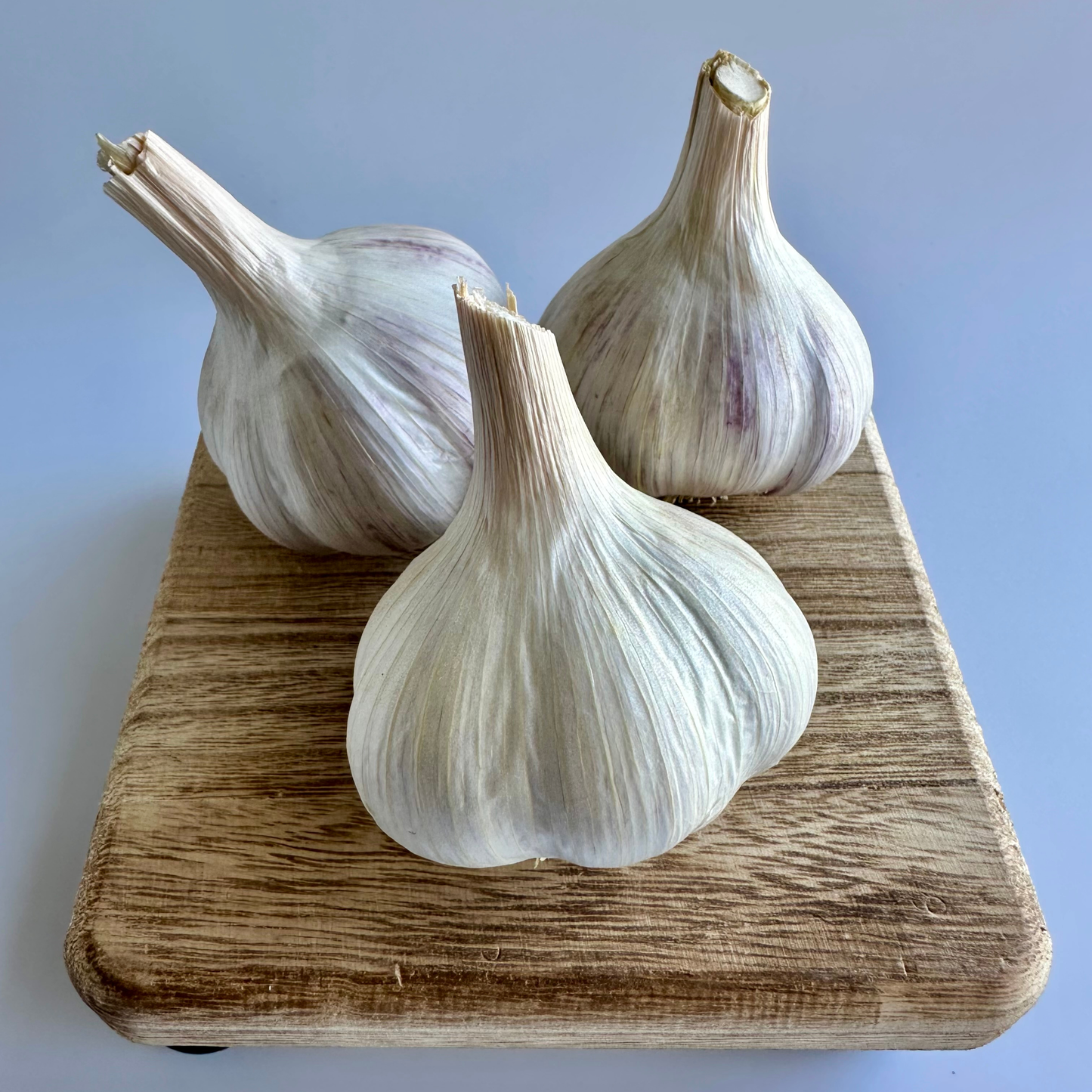 Three whole garlic bulbs with pale ivory-white to light beige skins and subtle purple streaks, arranged on a rectangular wooden cutting board against a white background.